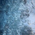 A tight school of silver sardines skimming a very pale sandy bottom dotted with dark rocks, the mass of fish densest toward the left, in the blue.