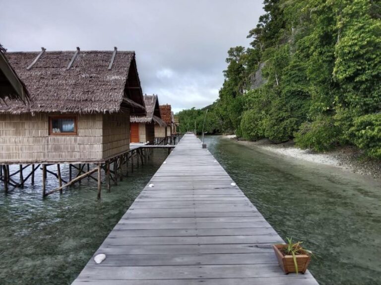A wooden walkway above the water, along the bungalows with palm-thatched roofs
