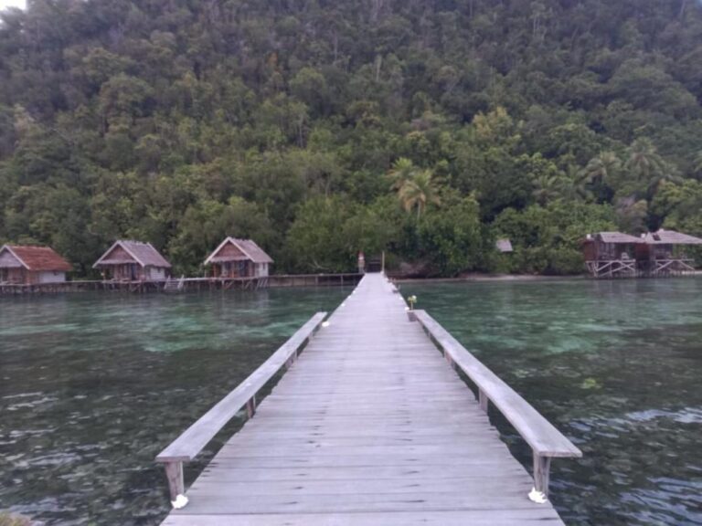 A long jetty with benches over calm water, facing the bungalows and the wooded hill