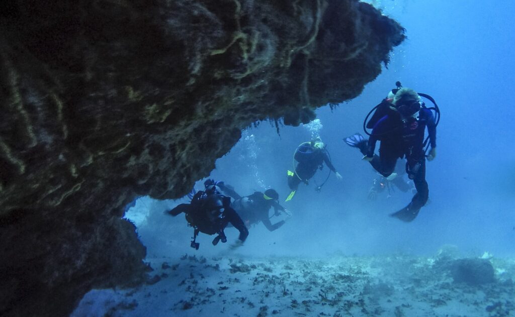 Four divers come out of a coral tunnel over a sandy bottom in Cozumel, the rocky ceiling above them