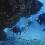 Four divers come out of a coral tunnel over a sandy bottom in Cozumel, the rocky ceiling above them