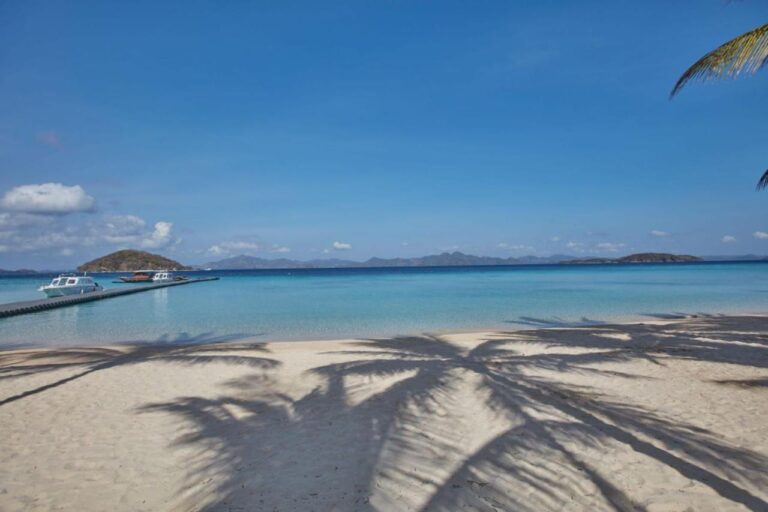 Palm tree shadows on the sand, facing the boats moored at the jetty and the islands in the distance