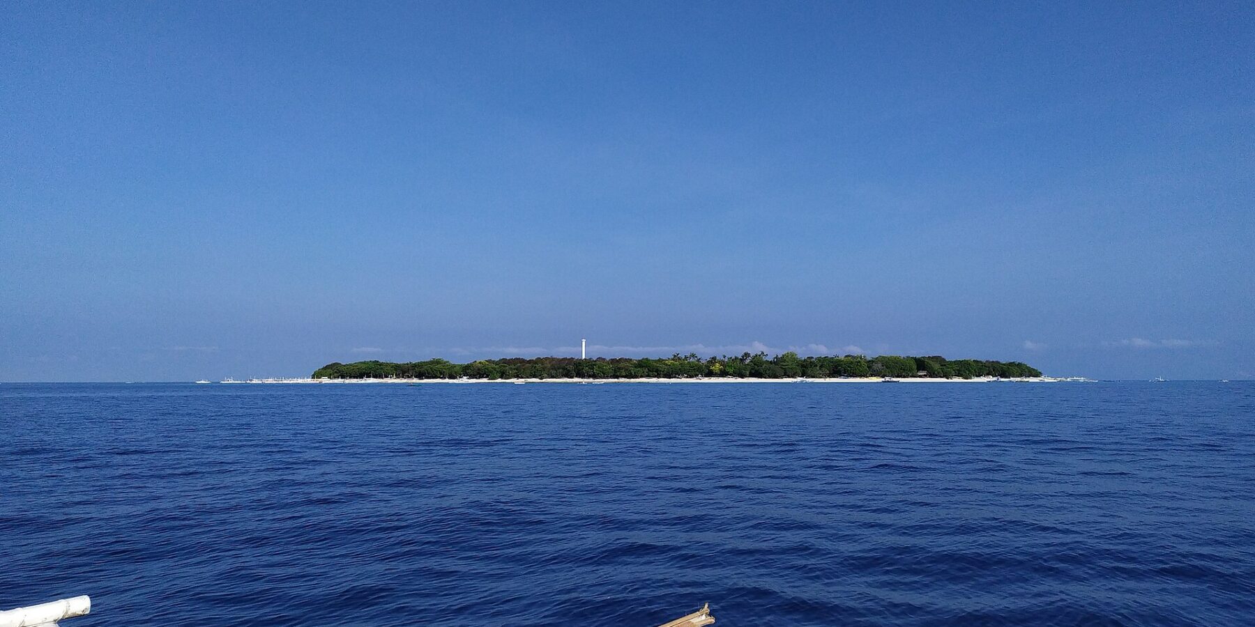 The flat, wooded island of Balicasag on the horizon, with its white lighthouse and fringe of pale sand, seen from a bangka whose white outriggers fill the foreground, under a cloudless blue sky.