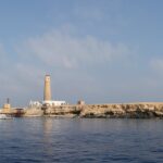 Big Brother from the sea: a flat, pale islet topped by its lighthouse, with a white liveaboard moored in front
