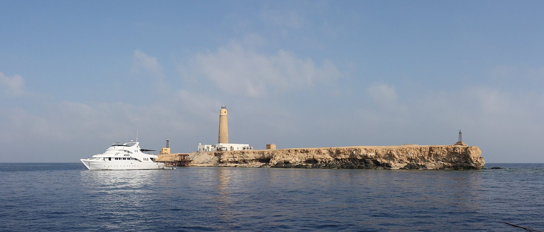 Big Brother from the sea: a flat, pale islet topped by its lighthouse, with a white liveaboard moored in front