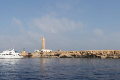Big Brother from the sea: a flat, pale islet topped by its lighthouse, with a white liveaboard moored in front