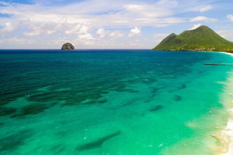 Aerial view of the long Le Diamant beach lined with turquoise water, Diamond Rock offshore and Morne Larcher, a green hill, on the right