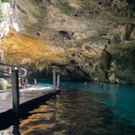 The east eye of Dos Ojos Cenote: a limestone vault bristling with stalactites above turquoise water, a wooden platform on the left and swimmers in the background