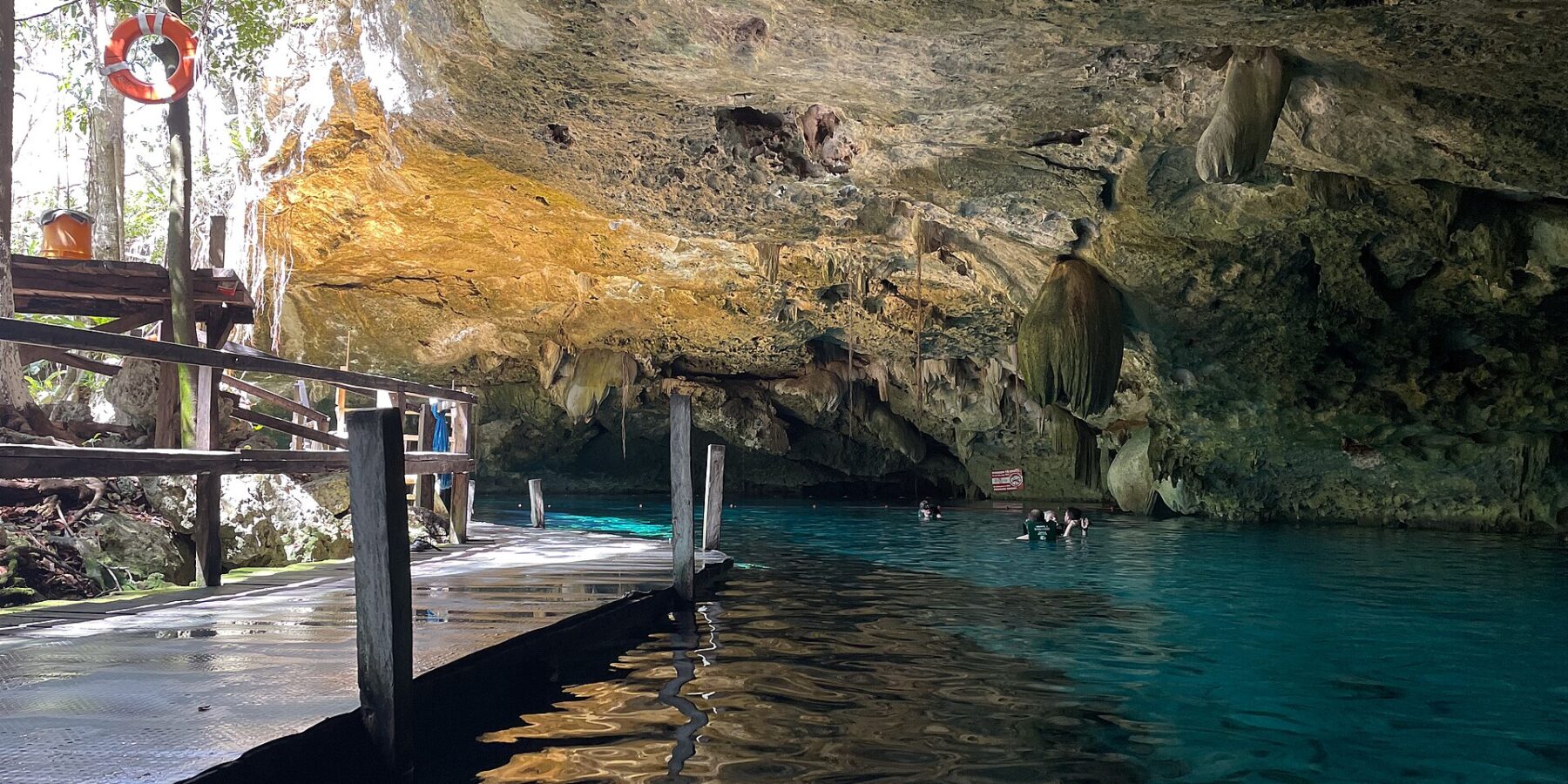 The east eye of Dos Ojos Cenote: a limestone vault bristling with stalactites above turquoise water, a wooden platform on the left and swimmers in the background
