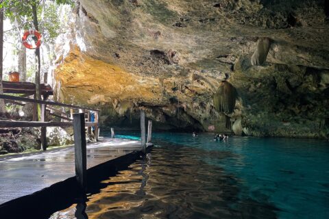 The east eye of Dos Ojos Cenote: a limestone vault bristling with stalactites above turquoise water, a wooden platform on the left and swimmers in the background
