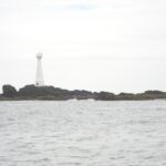 A slender white lighthouse with a red door stands on the low, rocky islet of Formigão, its dark rocks fringed with brown algae, above a gray, rough sea under an overcast sky