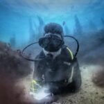A diver in a black wetsuit, lying on a sandy bottom between two rocks covered in brown algae, writes on a white slate, with the outlines of kelp standing out in the blue behind him.