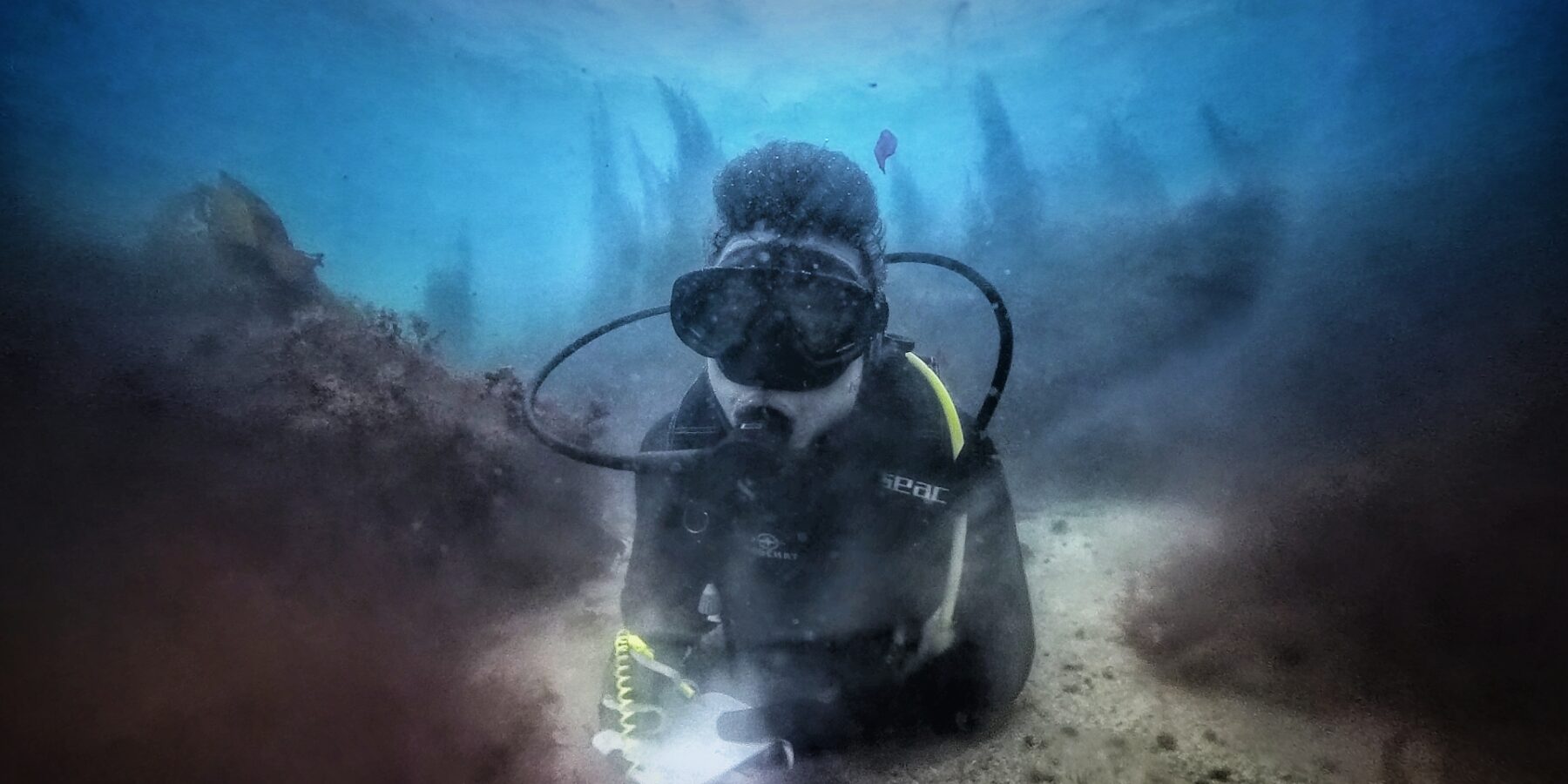 A diver in a black wetsuit, lying on a sandy bottom between two rocks covered in brown algae, writes on a white slate, with the outlines of kelp standing out in the blue behind him.