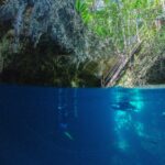 Split view at the entrance to The Pit Cenote: the limestone vault and wooden staircase under the jungle at the top, two divers in deep blue water below