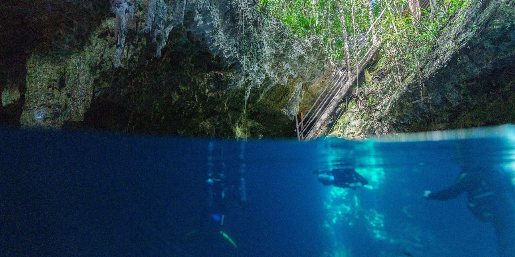 Split view at the entrance to The Pit Cenote: the limestone vault and wooden staircase under the jungle at the top, two divers in deep blue water below