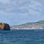 Panorama of the two ocher tuff islets of Madalena on a dark blue sea, the long flat Ilhéu Deitado on the left and the upright Ilhéu em Pé on the right, with the white town of Horta and the green hills of Faial behind