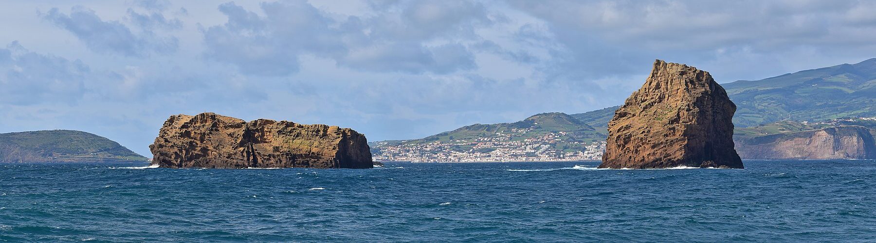 Panorama of the two ocher tuff islets of Madalena on a dark blue sea, the long flat Ilhéu Deitado on the left and the upright Ilhéu em Pé on the right, with the white town of Horta and the green hills of Faial behind