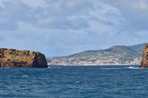 Panorama of the two ocher tuff islets of Madalena on a dark blue sea, the long flat Ilhéu Deitado on the left and the upright Ilhéu em Pé on the right, with the white town of Horta and the green hills of Faial behind