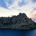 Maïre Island, a mass of pale limestone with jagged ridges, stands out against a sunset sky with pink and mauve clouds, above a dark blue sea.