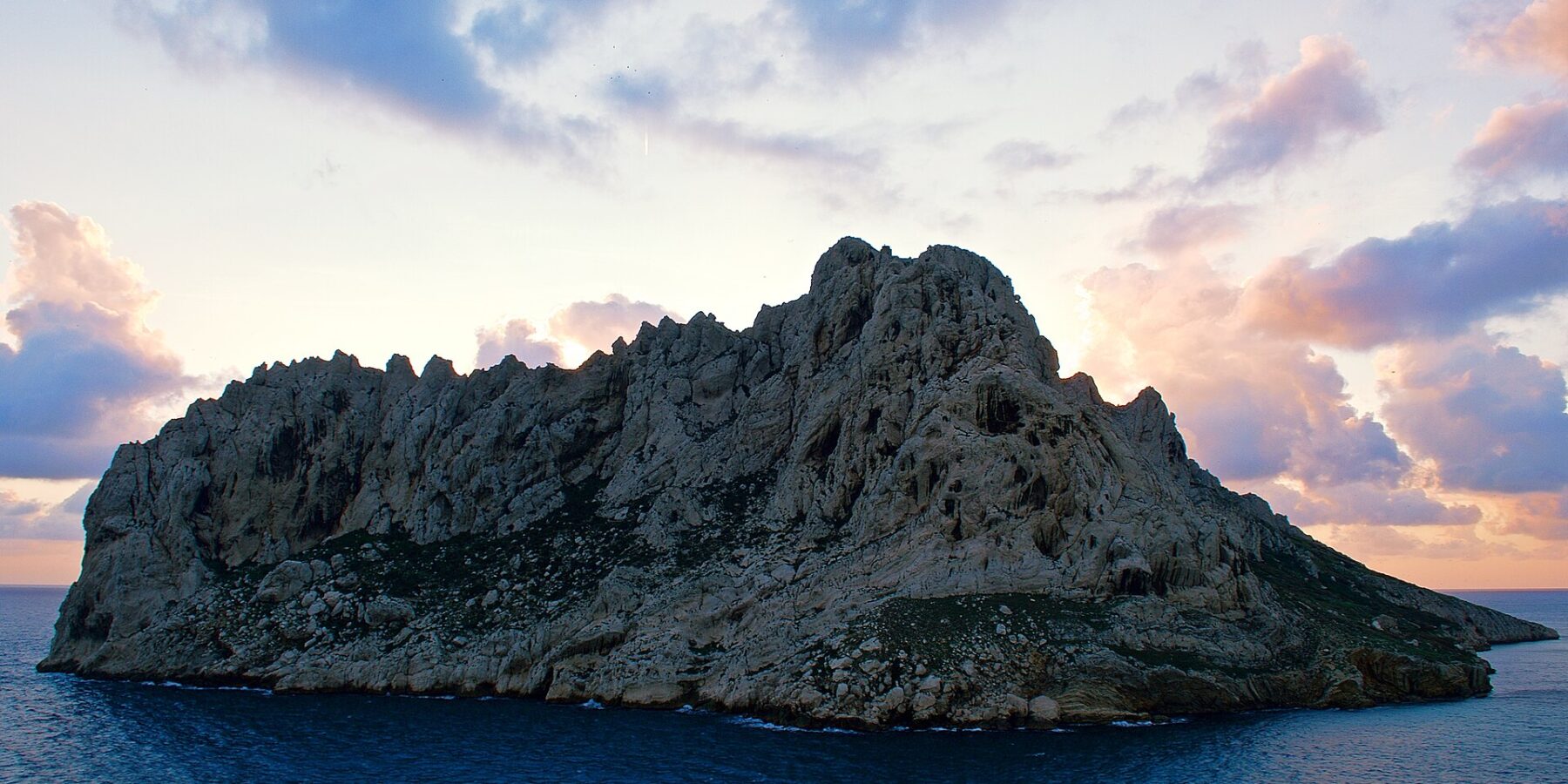 Maïre Island, a mass of pale limestone with jagged ridges, stands out against a sunset sky with pink and mauve clouds, above a dark blue sea.