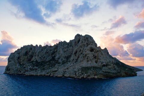 Maïre Island, a mass of pale limestone with jagged ridges, stands out against a sunset sky with pink and mauve clouds, above a dark blue sea.