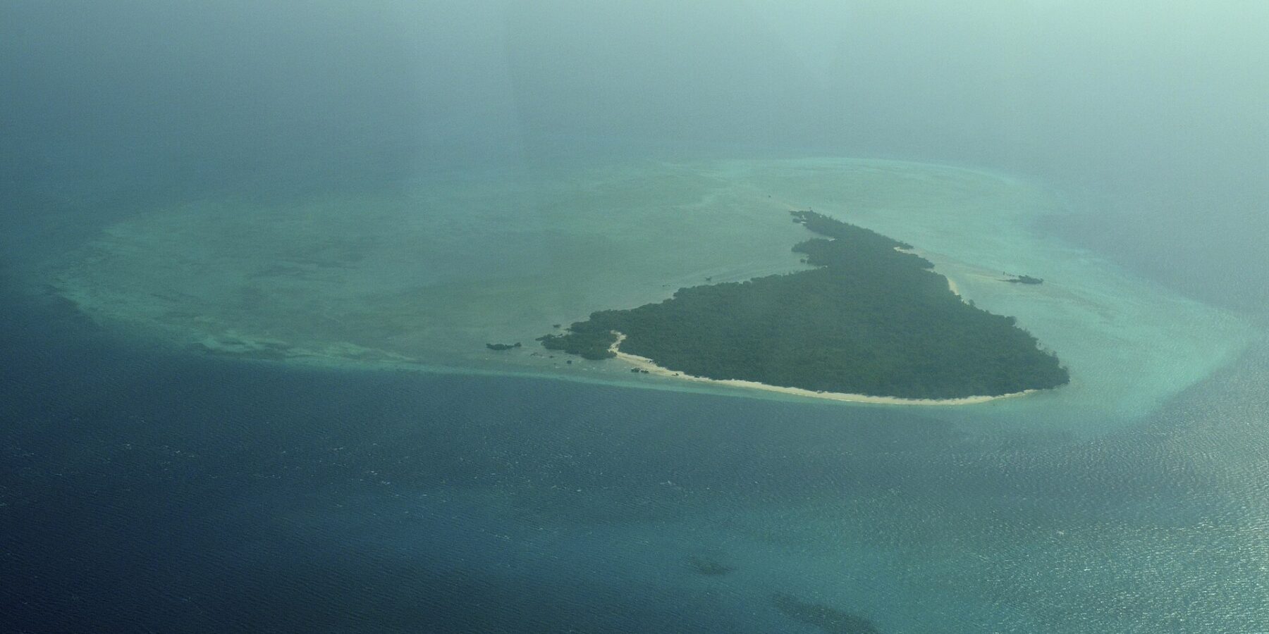 Misali Island seen from a plane, a triangle of forest edged by a white beach, sitting on a turquoise reef flat in a dark blue sea, in the haze