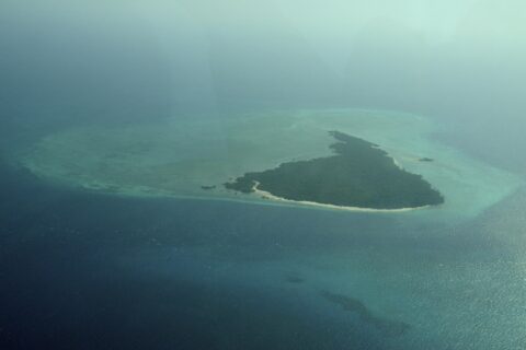 Misali Island seen from a plane, a triangle of forest edged by a white beach, sitting on a turquoise reef flat in a dark blue sea, in the haze