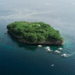 Aerial view of Pescador Island, a rock covered in green vegetation with its white lighthouse, surrounded by about ten dive bancas on a dark blue sea