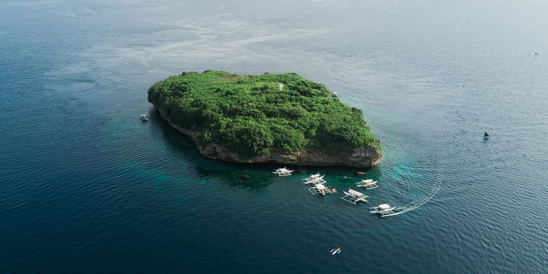 Aerial view of Pescador Island, a rock covered in green vegetation with its white lighthouse, surrounded by about ten dive bancas on a dark blue sea