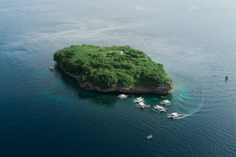Aerial view of Pescador Island, a rock covered in green vegetation with its white lighthouse, surrounded by about ten dive bancas on a dark blue sea