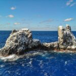 Drone view of Roca Partida: a bird-whitened rock split into two peaks, pounded by a deep blue sea under a blue sky dotted with clouds