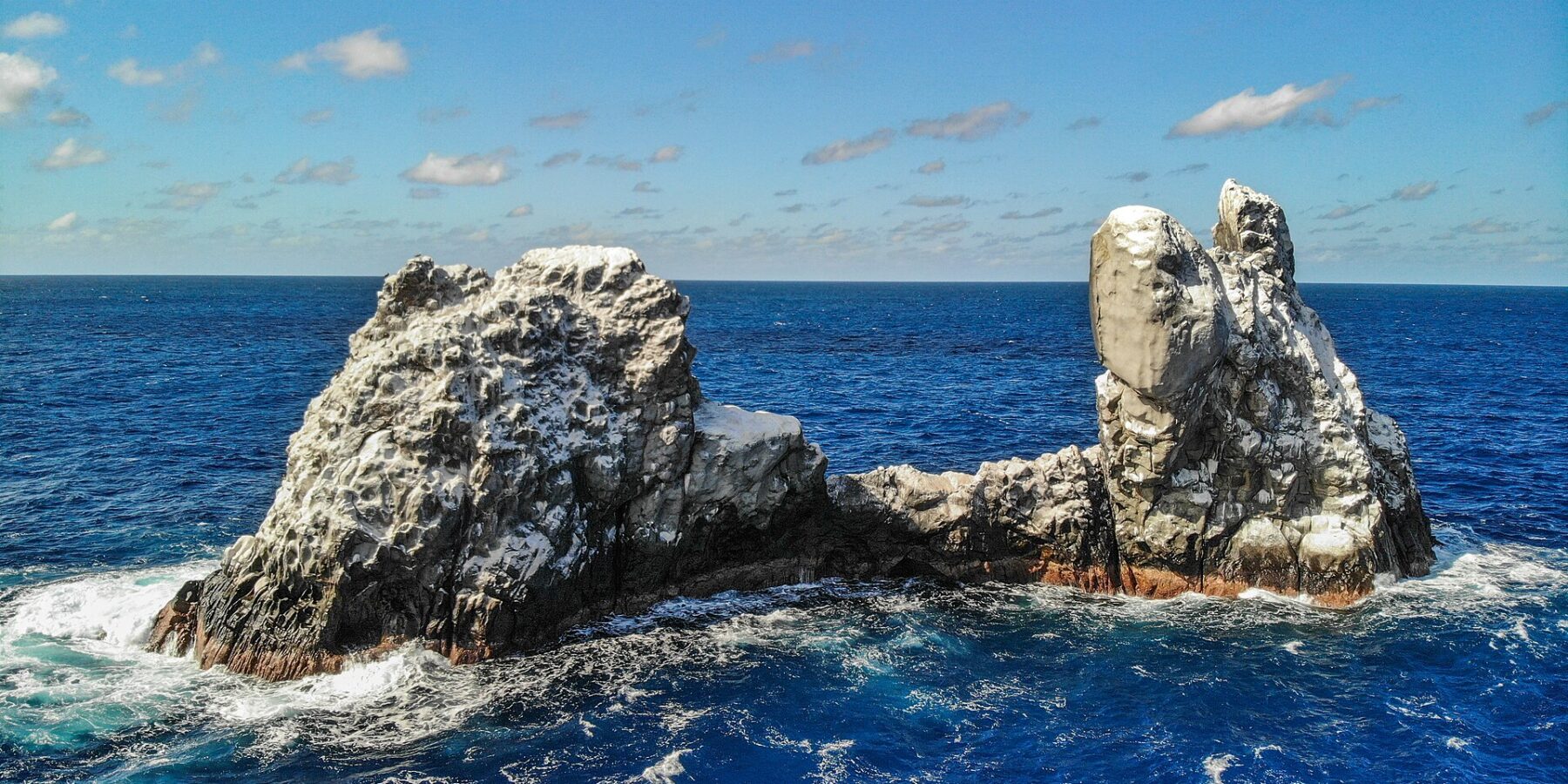 Drone view of Roca Partida: a bird-whitened rock split into two peaks, pounded by a deep blue sea under a blue sky dotted with clouds