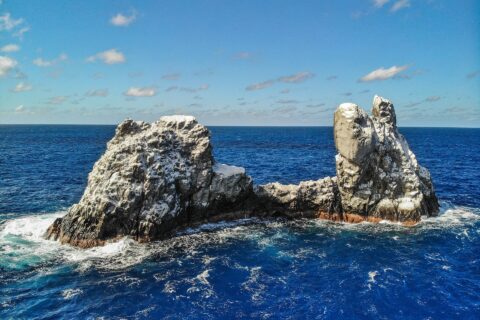 Drone view of Roca Partida: a bird-whitened rock split into two peaks, pounded by a deep blue sea under a blue sky dotted with clouds