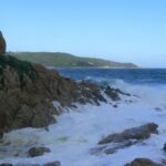 Waves break into white foam on the granite rocks of L'Escalet in the foreground, facing a rough dark blue sea, with the wooded coast of Cap Camarat and its white lighthouse in the distance.