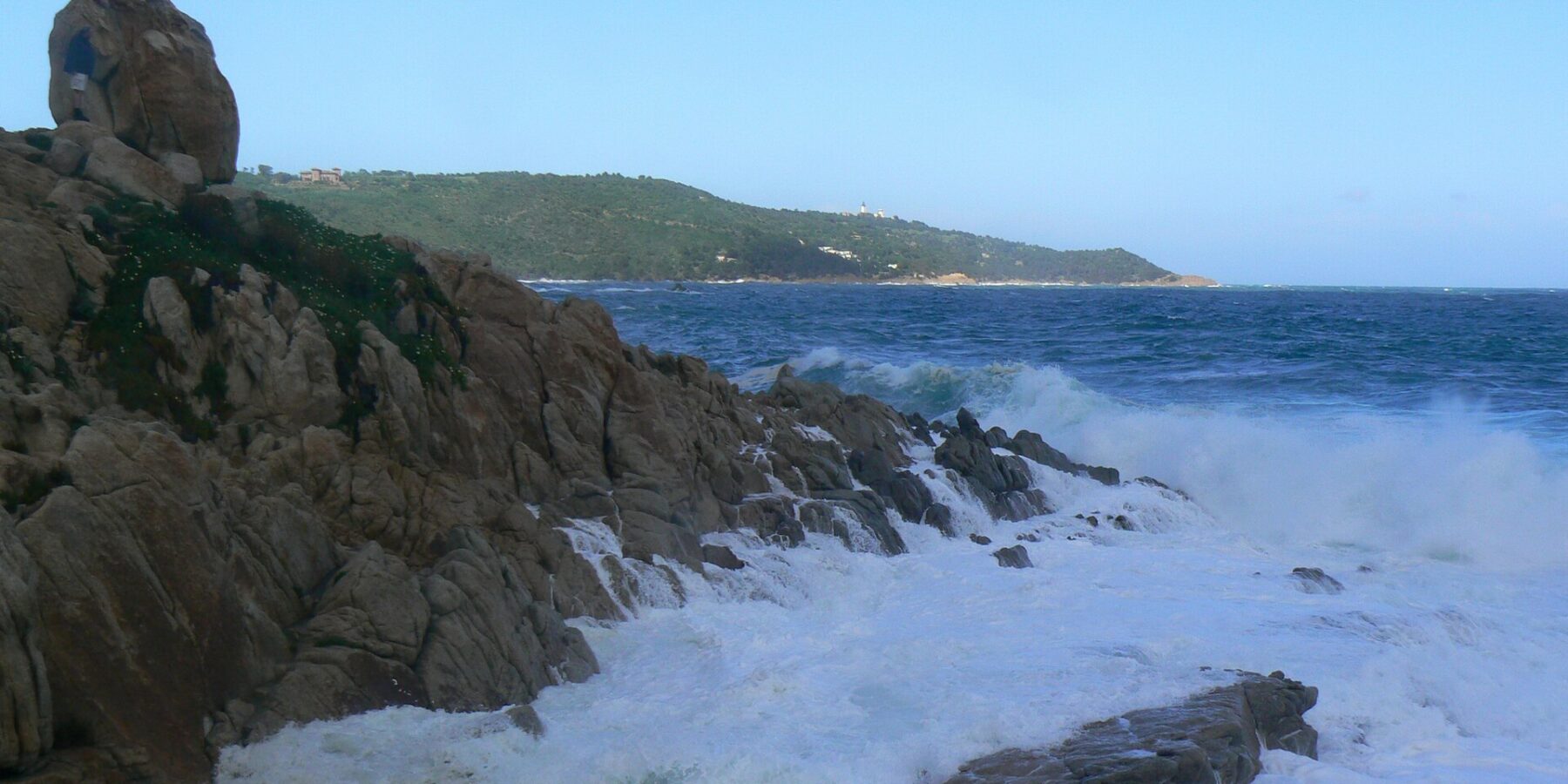 Waves break into white foam on the granite rocks of L'Escalet in the foreground, facing a rough dark blue sea, with the wooded coast of Cap Camarat and its white lighthouse in the distance.