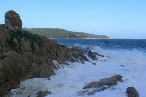 Waves break into white foam on the granite rocks of L'Escalet in the foreground, facing a rough dark blue sea, with the wooded coast of Cap Camarat and its white lighthouse in the distance.