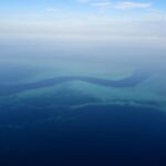 Aerial view of a dark blue S-shaped channel winding between the turquoise flats of the barrier reef, with a few small white boats in the pass