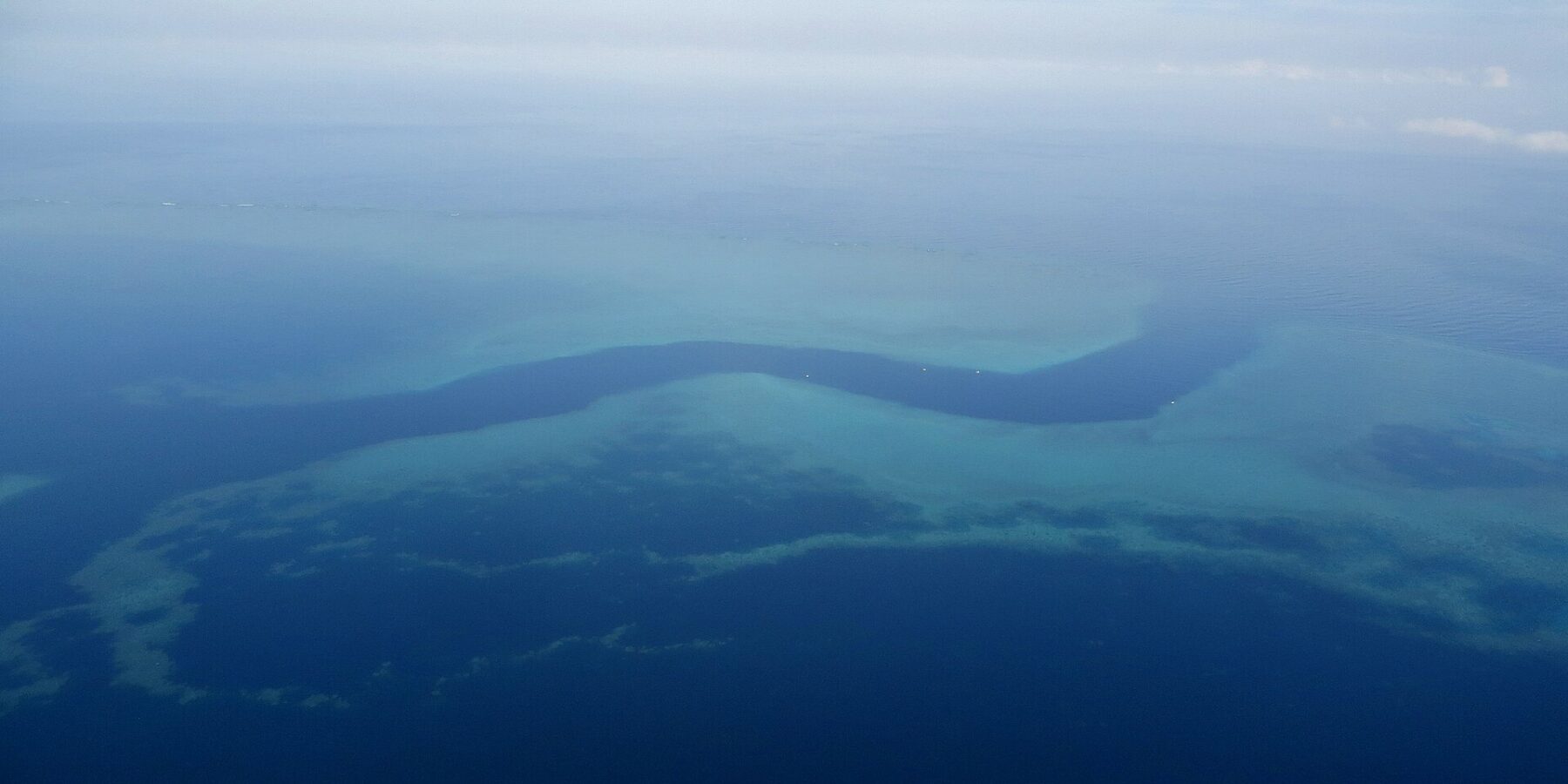 Aerial view of a dark blue S-shaped channel winding between the turquoise flats of the barrier reef, with a few small white boats in the pass
