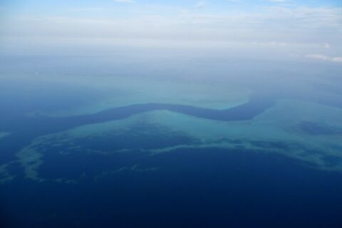 Aerial view of a dark blue S-shaped channel winding between the turquoise flats of the barrier reef, with a few small white boats in the pass