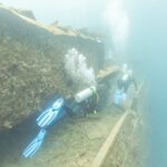 Two divers swim along the deck of the Thistlegorm beside a passageway, in milky blue water