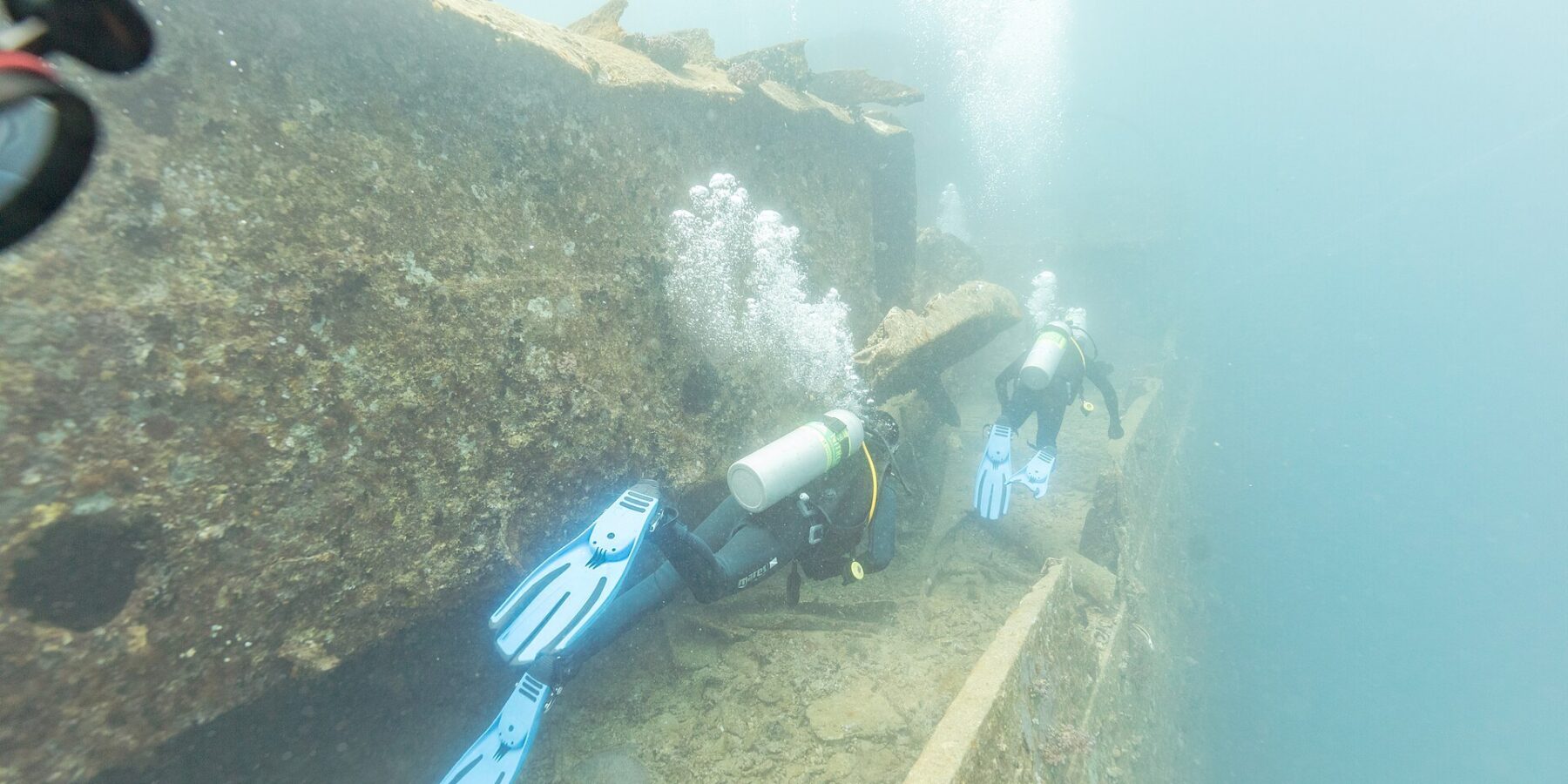 Two divers swim along the deck of the Thistlegorm beside a passageway, in milky blue water