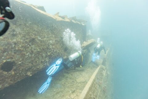 Two divers swim along the deck of the Thistlegorm beside a passageway, in milky blue water