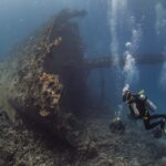 The upright bow of the Giannis D, covered in growth, with three divers at the foot of the wreck in blue water full of bubbles