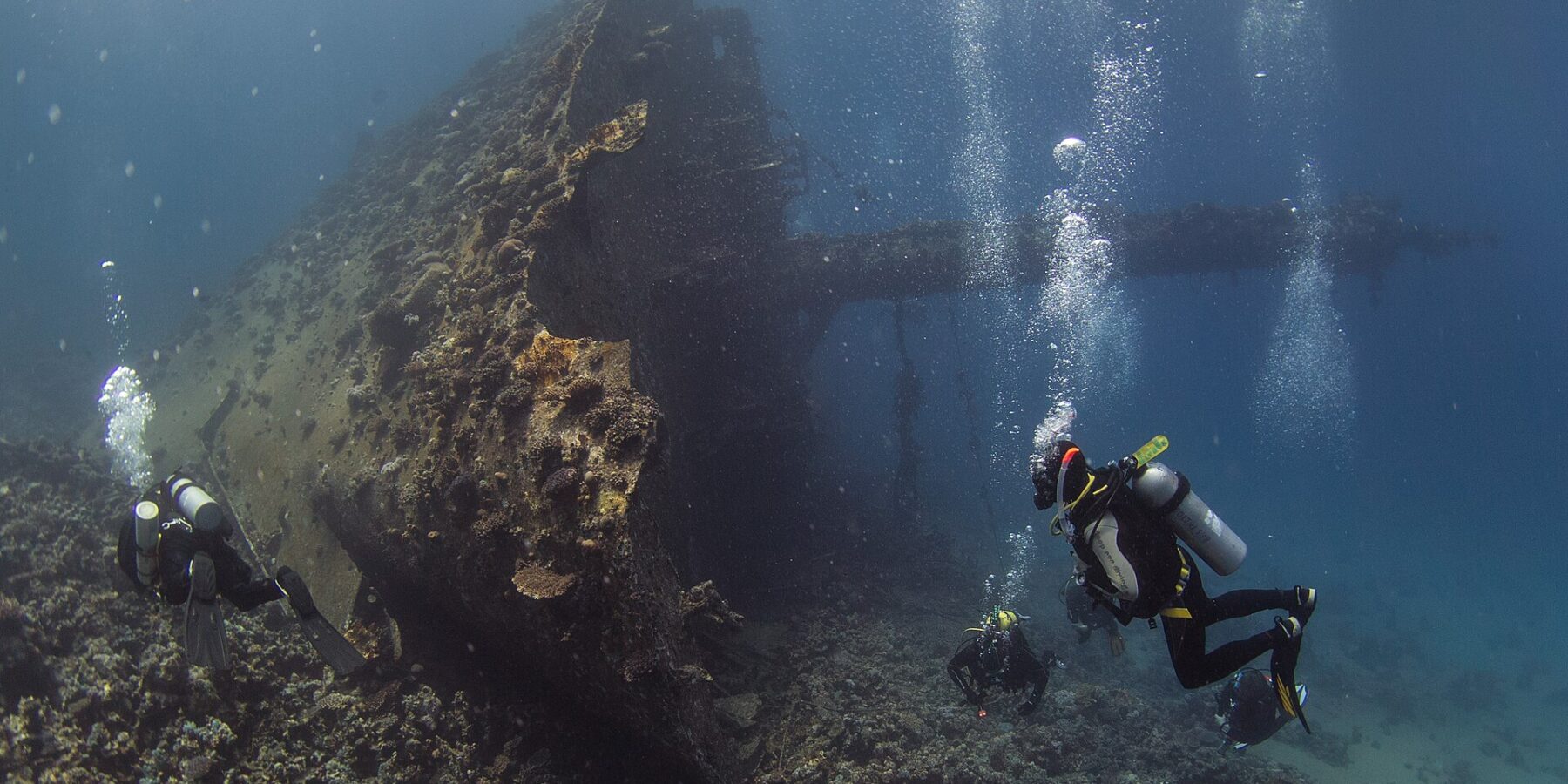 The upright bow of the Giannis D, covered in growth, with three divers at the foot of the wreck in blue water full of bubbles