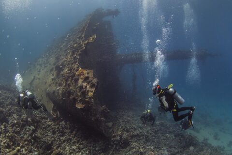 The upright bow of the Giannis D, covered in growth, with three divers at the foot of the wreck in blue water full of bubbles