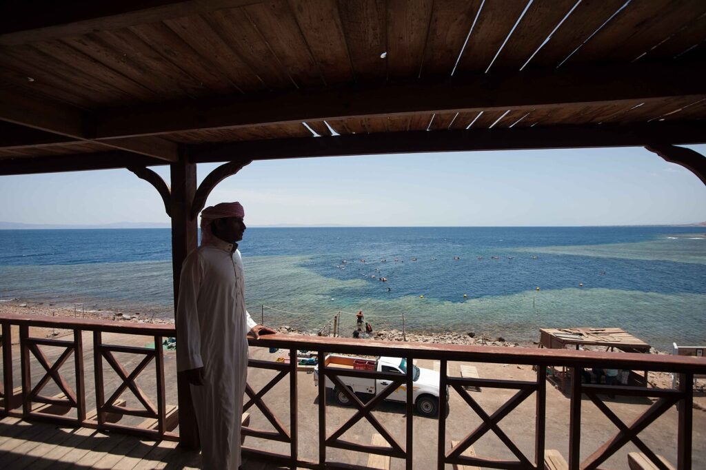 From a wooden terrace, a man in a galabeya looks at the Blue Hole, ringed by pale reef, where people are swimming