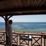 From a wooden terrace, a man in a galabeya looks at the Blue Hole, ringed by pale reef, where people are swimming
