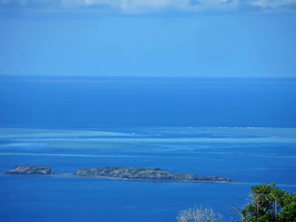 View looking down on the blue lagoon, two rocky islets in the foreground and, in the distance, the pale flats of the barrier reef where a white boat sits in the pass