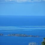 View looking down on the blue lagoon, two rocky islets in the foreground and, in the distance, the pale flats of the barrier reef where a white boat sits in the pass