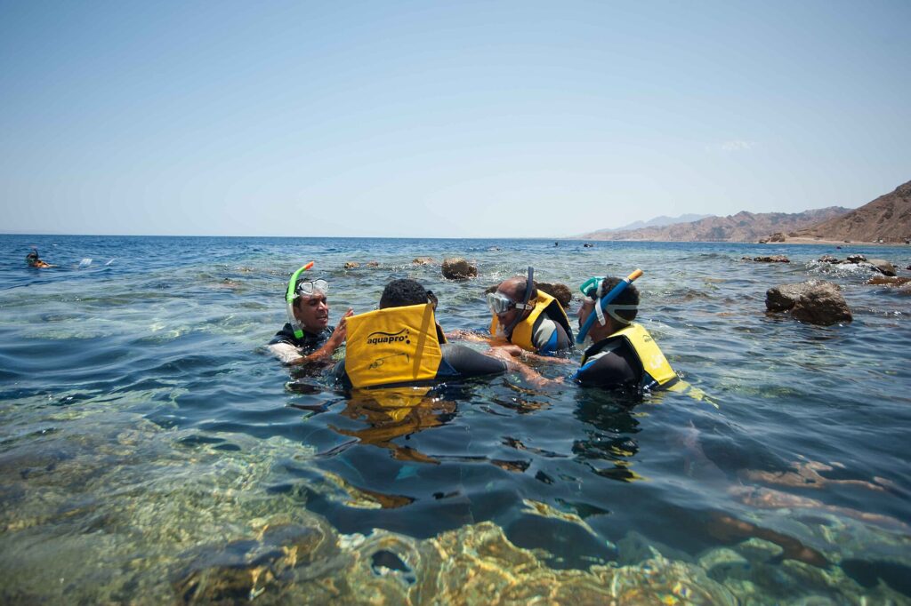 Swimmers in yellow vests, masks and snorkels in the clear water of the Blue Hole, with the Sinai mountains behind